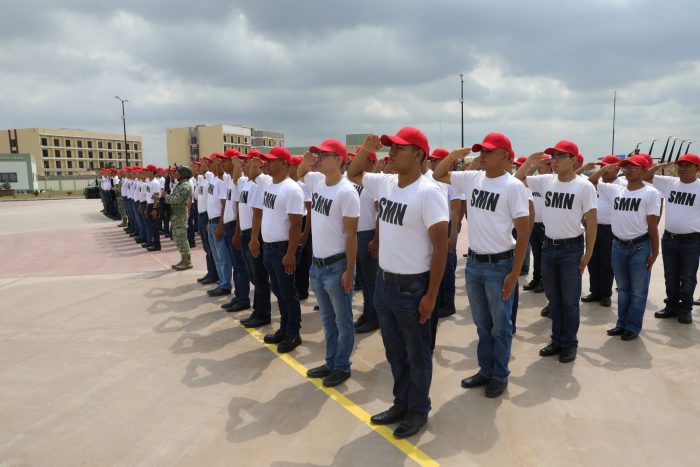 Line of recruits in white SMN shirts and red caps standing at attention and saluting on a concrete outdoor area, with a drill instructor in camouflage nearby.