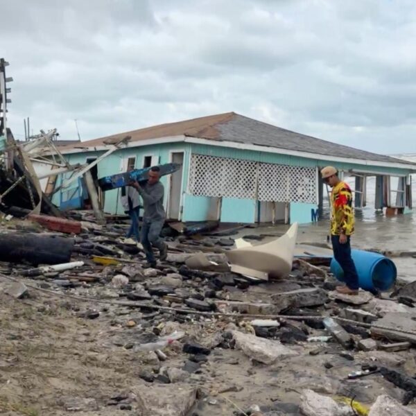 Men salvage debris from a collapsed seaside house after a storm, along a rocky, debris-strewn shore with rough waves in the background.