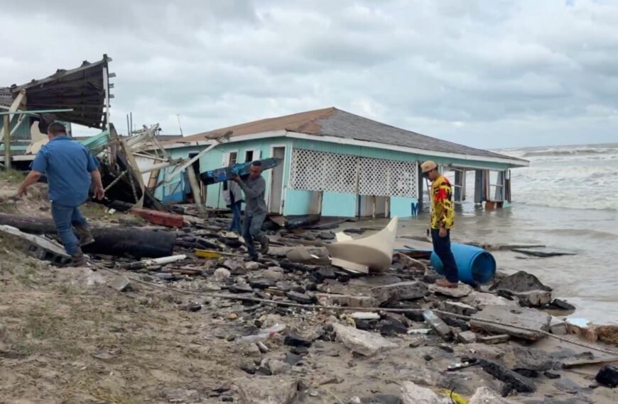Men salvage debris from a collapsed seaside house after a storm, along a rocky, debris-strewn shore with rough waves in the background.