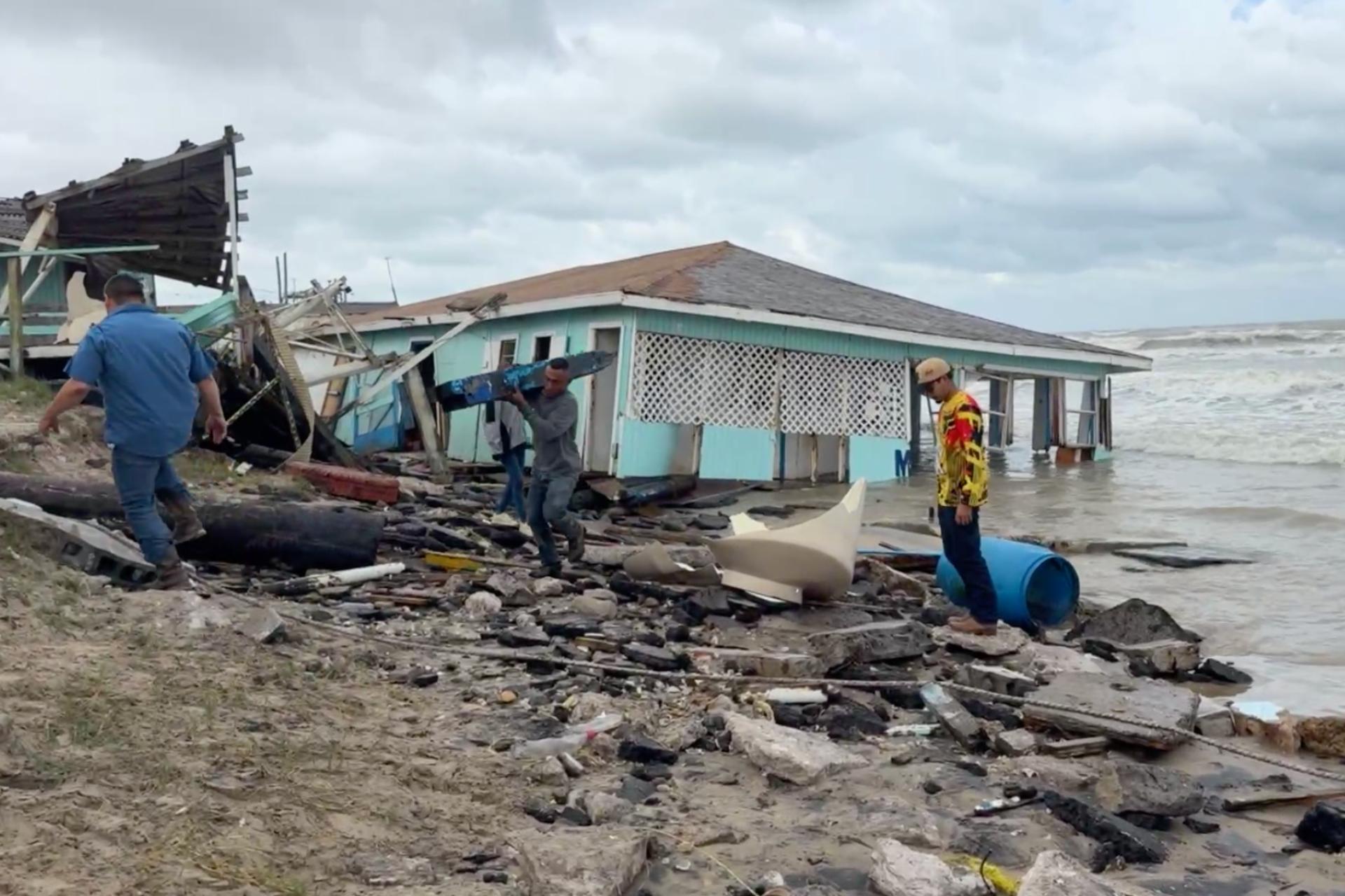 Men salvage debris from a collapsed seaside house after a storm, along a rocky, debris-strewn shore with rough waves in the background.