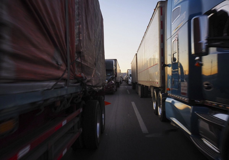 Line of large trucks riding side by side on a highway at sunset, with a blue truck on the right and a red-tarped trailer on the left.