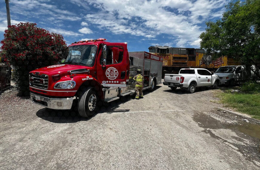 Red fire truck with a firefighter in full turnout gear standing beside it in a gravel lot, a white pickup nearby and a yellow train engine in the background.