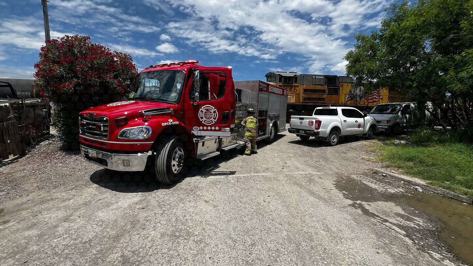 Red fire truck with a firefighter in full turnout gear standing beside it in a gravel lot, a white pickup nearby and a yellow train engine in the background.