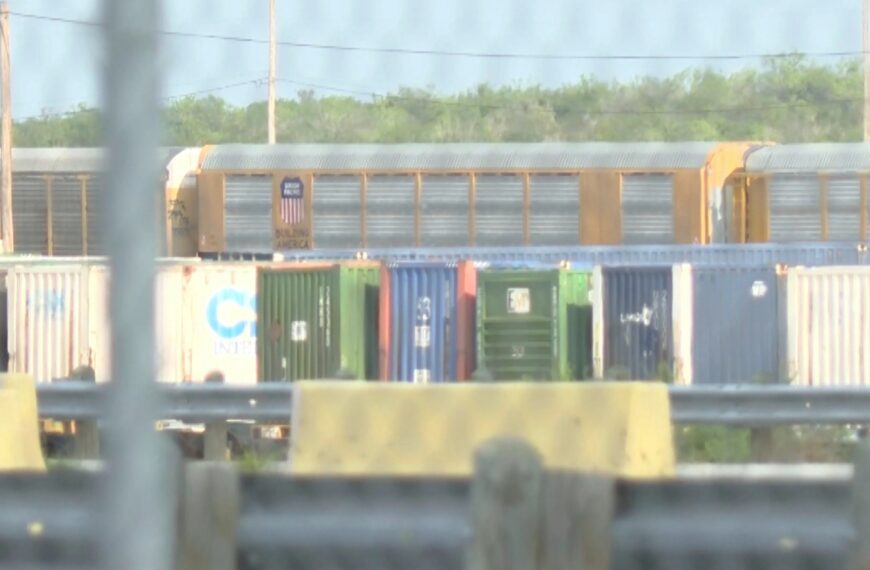 Blurry view of colorful freight cars in a rail yard behind a fence and barriers, with greenery in the background