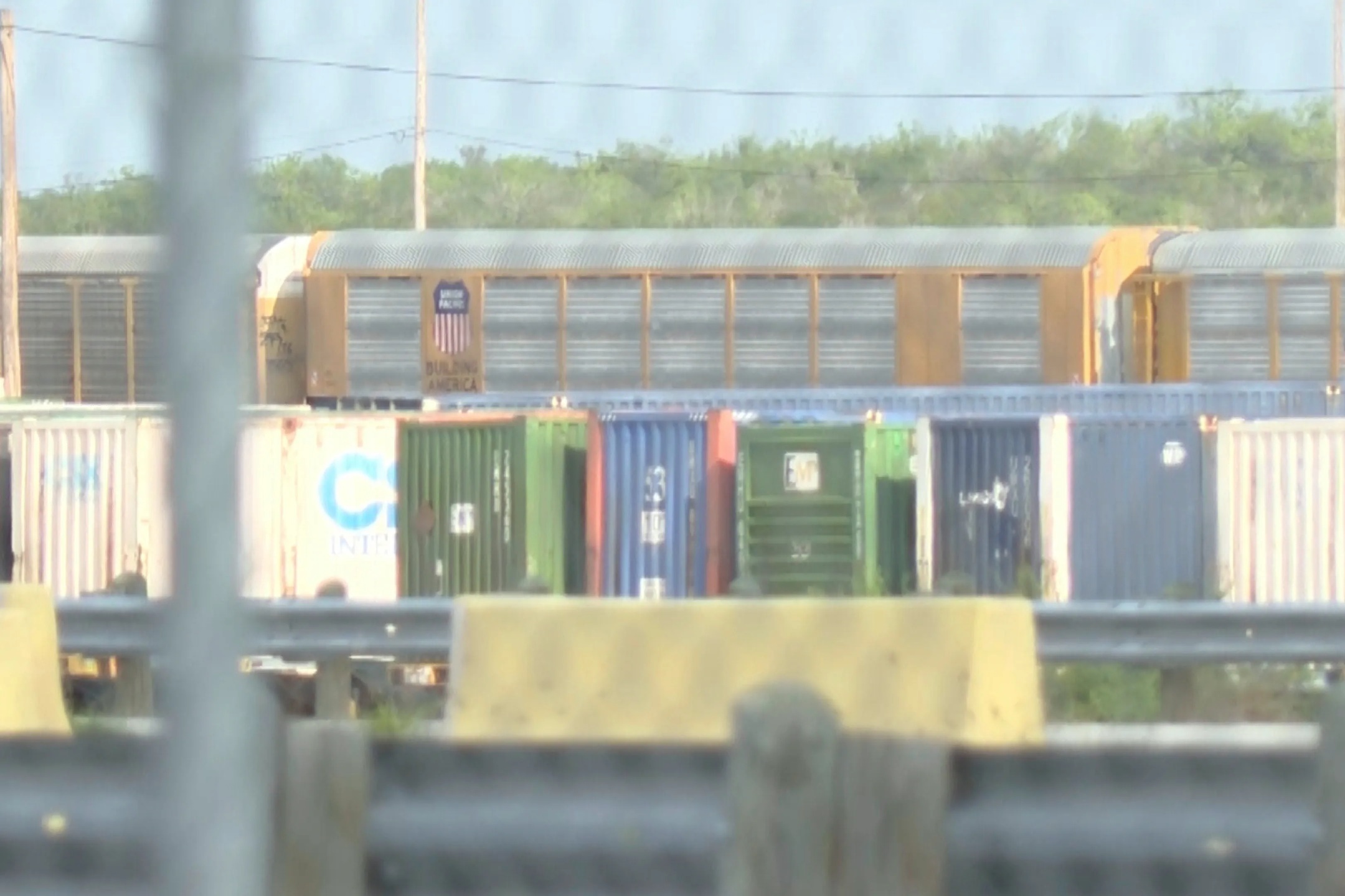Blurry view of colorful freight cars in a rail yard behind a fence and barriers, with greenery in the background