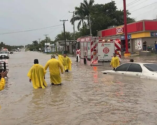 Rescuers in bright yellow ponchos wade through knee‑to‑waist deep floodwater past an overturned car and a parked fire truck near a storefront; onlookers photograph the scene.