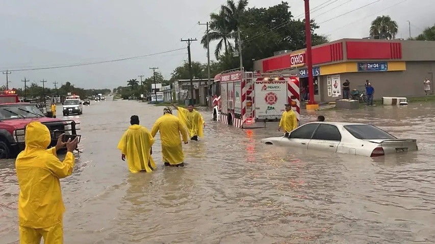 Rescuers in bright yellow ponchos wade through knee‑to‑waist deep floodwater past an overturned car and a parked fire truck near a storefront; onlookers photograph the scene.