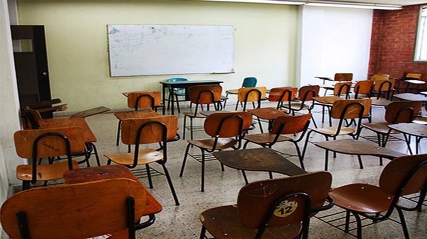 Empty classroom with wooden desks and chairs arranged in rows, a whiteboard at the front, and a few items on a teacher's desk.