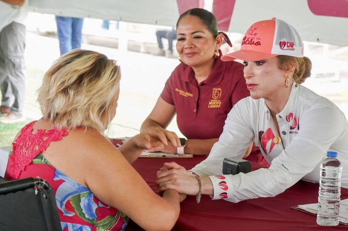 Volunteer in a white cap and long-sleeve shirt holds an older woman's hand across a table at an outreach event; another smiling volunteer in a maroon shirt watches nearby.