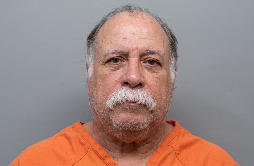 Close-up of an older man with a gray mustache and sparse hair wearing an orange shirt against a gray studio backdrop, looking straight at the camera.